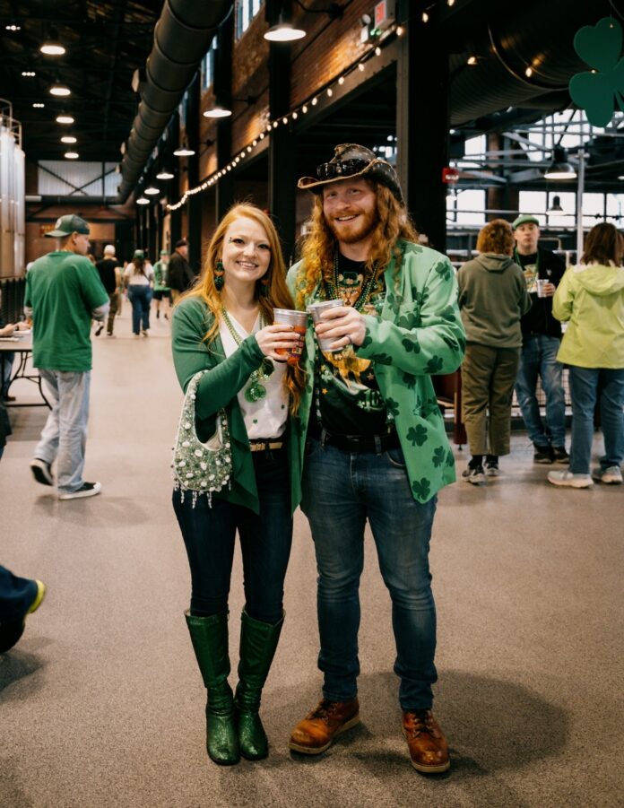 A man and a woman in St. Patrick's Day green clothing with shamrocks everywhere cheers a beer with their arms around each other.