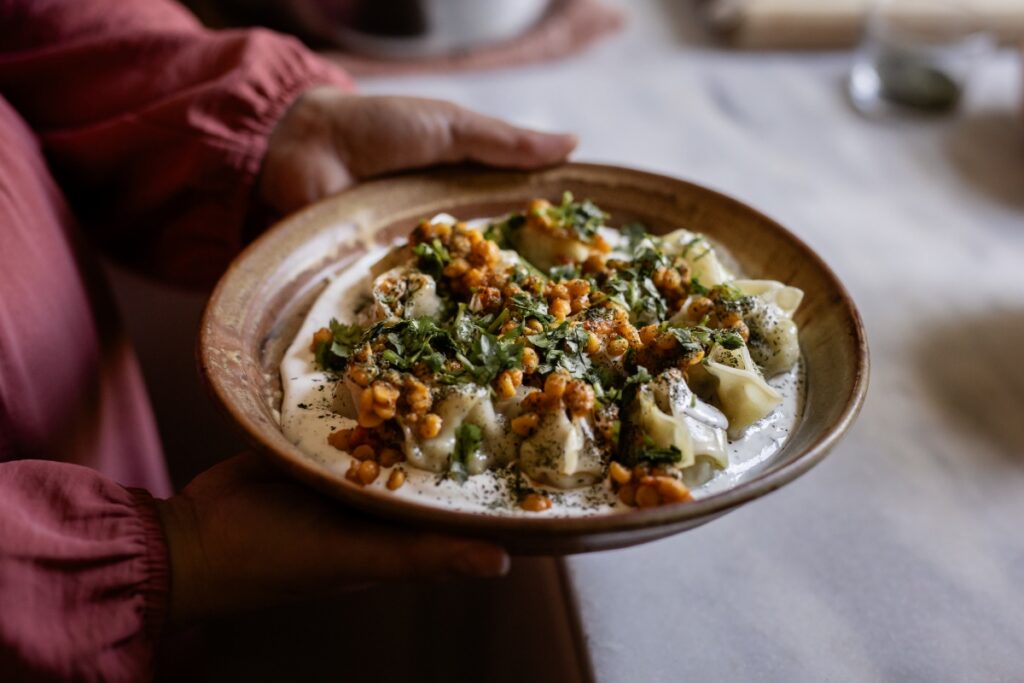 A woman wearing a black hijab and pink dress carries a large platter of traditional Afghan food through a kitchen with neutral-toned cabinetry.