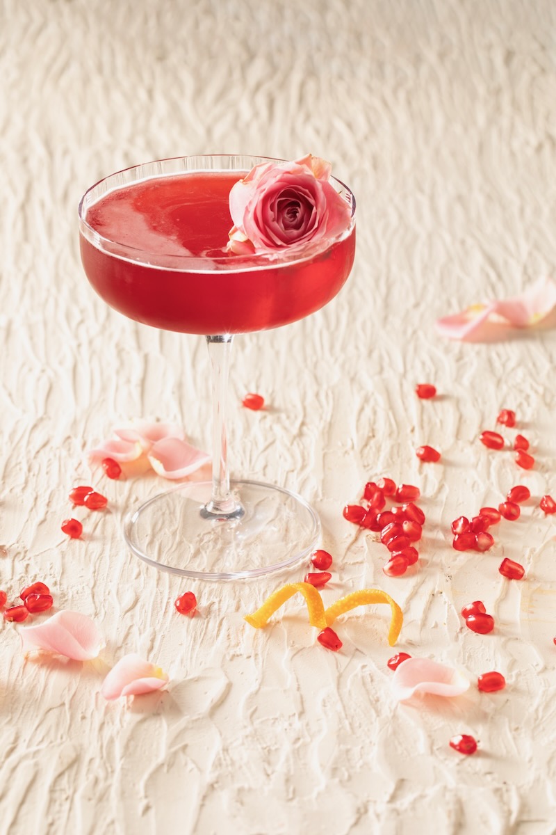 A red pomegranate cosmo as a Pretty Woman inspired drink sits in a tall martini glass with an edible rose floating on top and pomegranate seeds and an orange twist laying on the white table below.