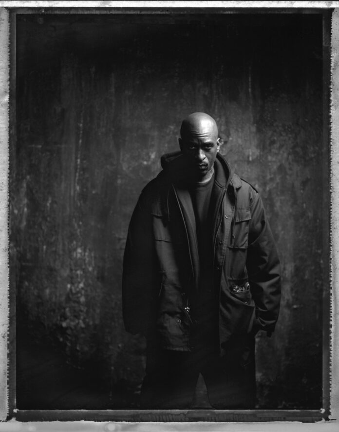 A man stands in a big puffy jacket against a black wall.