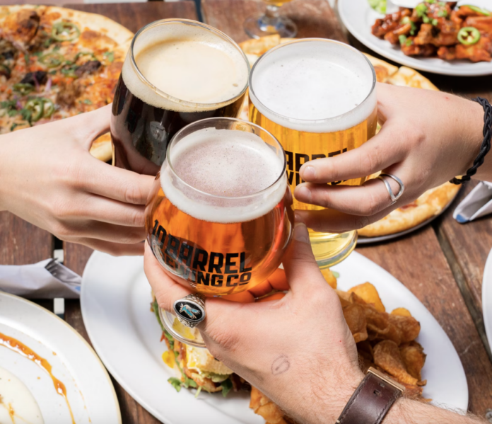 Three people cheers three glasses of beer in the center of a spead of foods like pizza, burgers, chips.