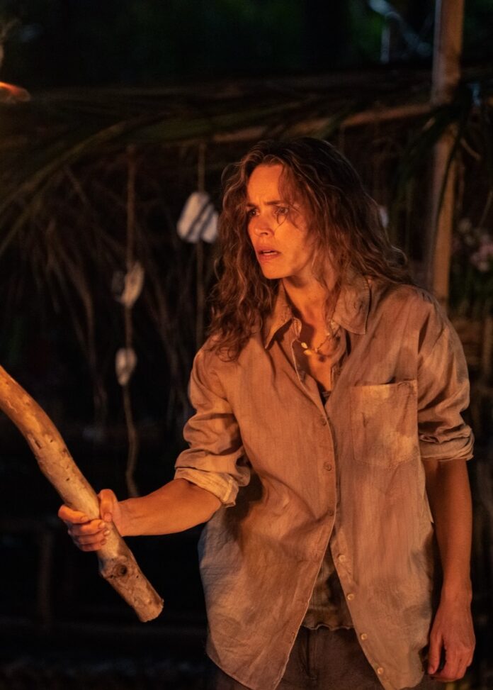 A woman stands holding a large stick in the firelight.