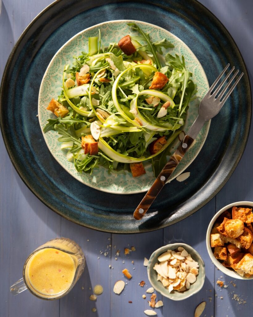 A plate full of shaved asparagus and croutons with a salad dressing container in the bottom left corner.