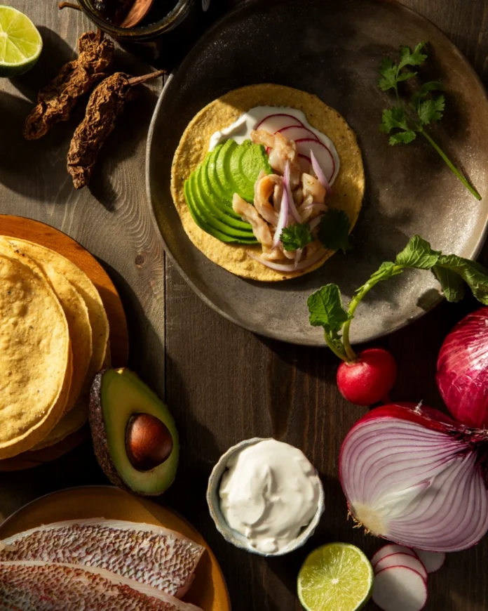 A fresh red snapper ceviche tostada topped with sliced avocado, radishes, and red onion, surrounded by raw ingredients like limes, dried chipotle peppers, and snapper filets.