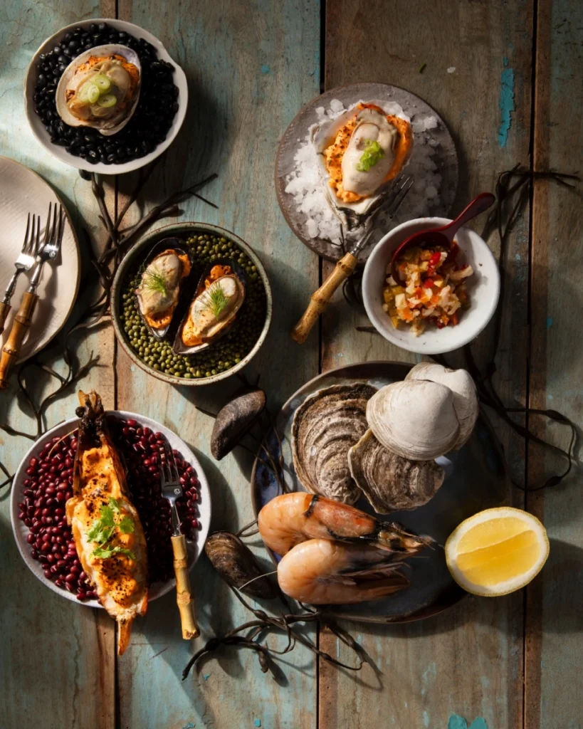 A rustic seafood platter featuring steamed prawns, mussels, and clams topped with Spicy Giardiniera Pimento Cheese, served on beds of colorful legumes and garnished with fresh herbs and lemon.