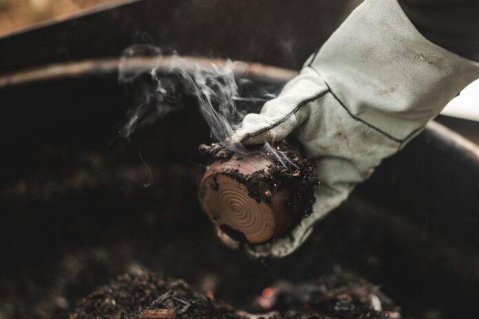 A gloved hand pulls a still-smoking, primitive-fired ceramic pot from a pit of glowing coals after an overnight outdoor firing session.
