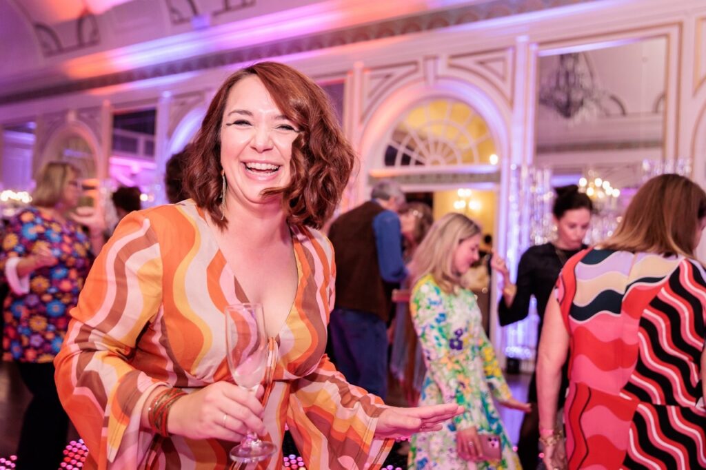 A woman in an orange and yellow and pink dress smiles with a champagne glass in hand.