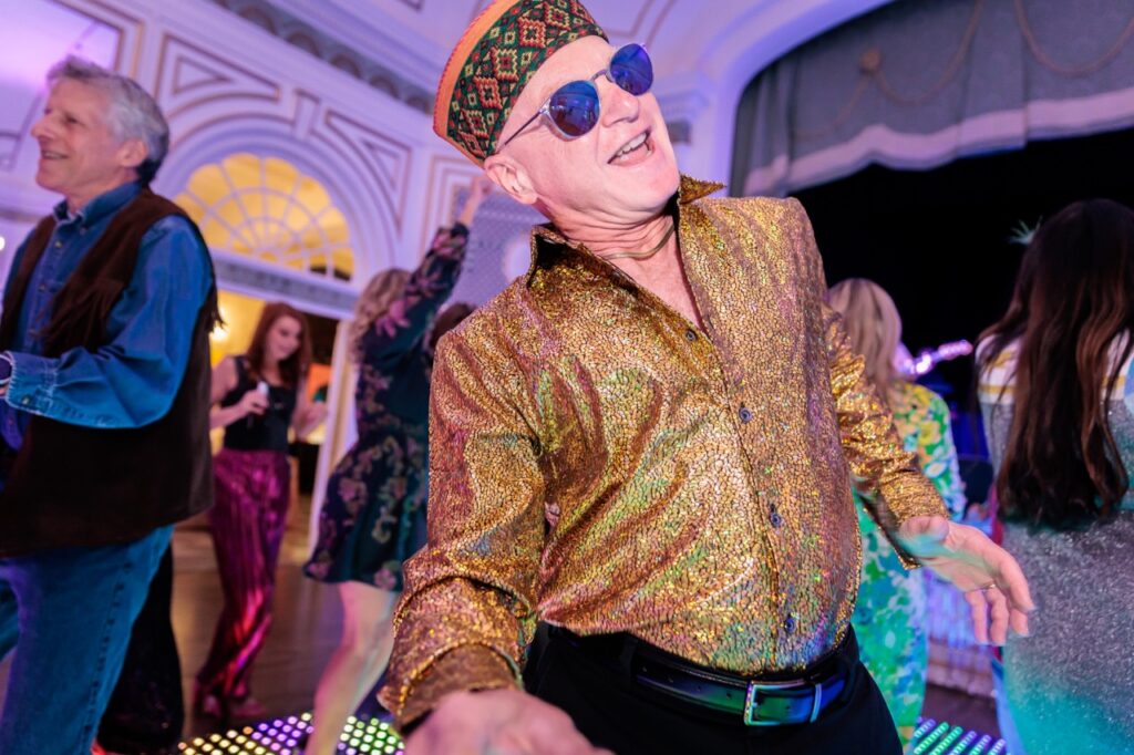 A man in a gold shirt and sunglasses dances on the dance floor.