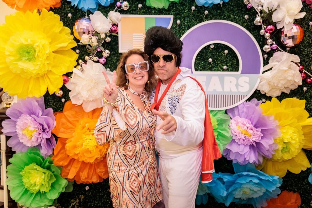 A man as Elvis and a woman in a pattern orange dress and large sunglasses stand in front of large flowers.