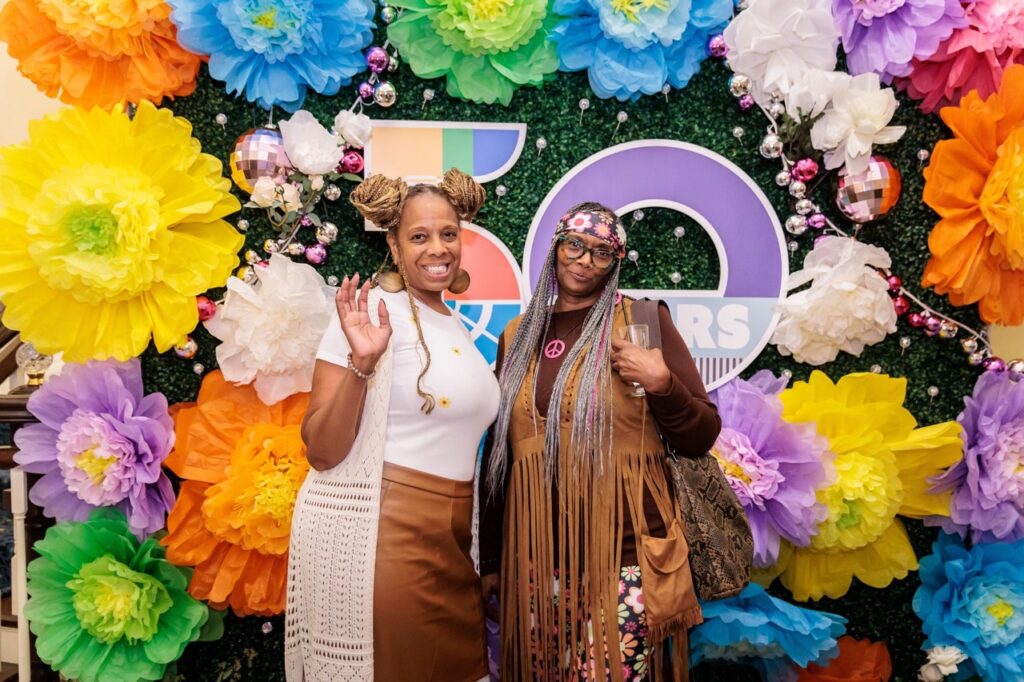 Two women in hippie outfits stand in front of a wall of paper flowers.