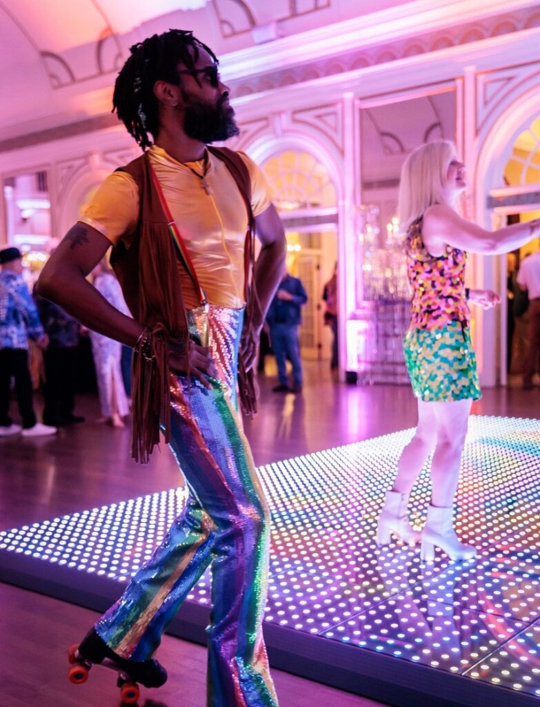 A man in a disco outfit roller skating on a wood floor.