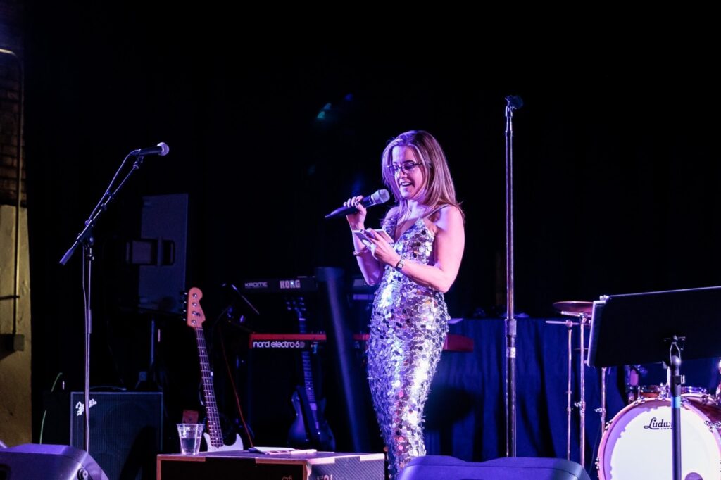 A woman in a sparkly dress stands in stage holding her phone.