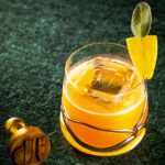 A bright yellow Teacher's Pet cocktail with apple brandy and sage honey, garnished with a lemon peel and sage leaf in a unique glass.
