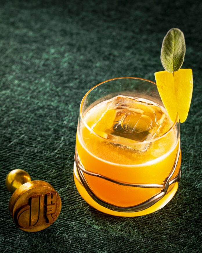 Teacher's Pet Cocktail TABLE Magazine A bright yellow Teacher's Pet cocktail with apple brandy and sage honey, garnished with a lemon peel and sage leaf in a unique glass.