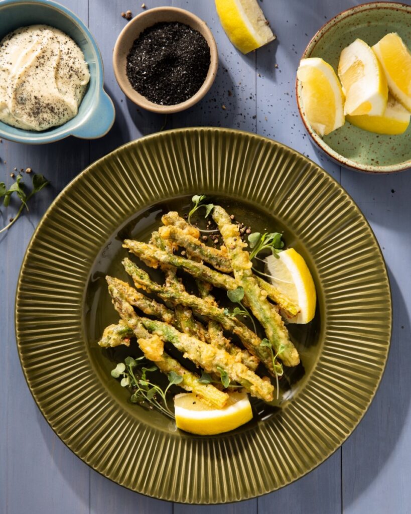 A green plate of lightly fried asparagus tempura with slices of lemon around it.