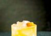 The Terms of My Surrender (Cinnamon-Spiced Rye & Rum Cocktail) A vibrant golden drink served over ice in a textured glass, garnished with an orange round and featuring rye and rum cocktail.