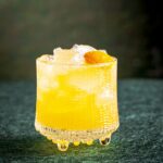 A vibrant golden drink served over ice in a textured glass, garnished with an orange round and featuring rye and rum cocktail.