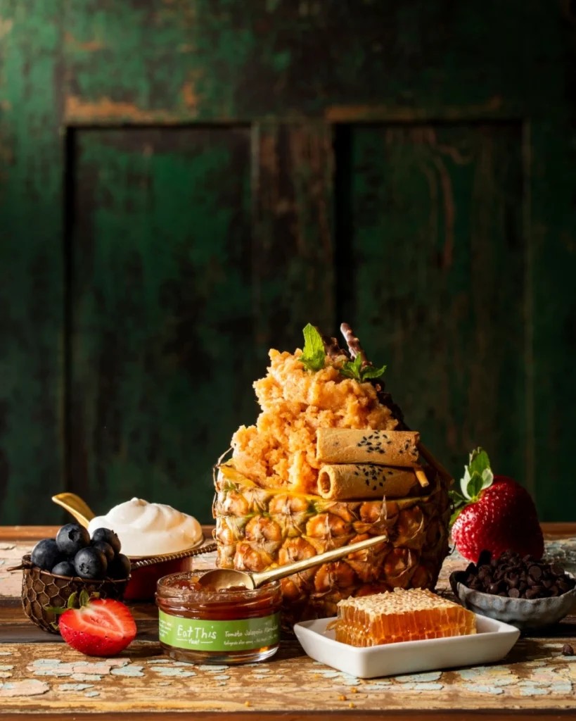 A vibrant Tomato Jalapeño Marmalade and Pineapple Granita served in a hollowed-out pineapple, garnished with chocolate-covered pocky and mint, and surrounded by fresh berries and honeycomb.