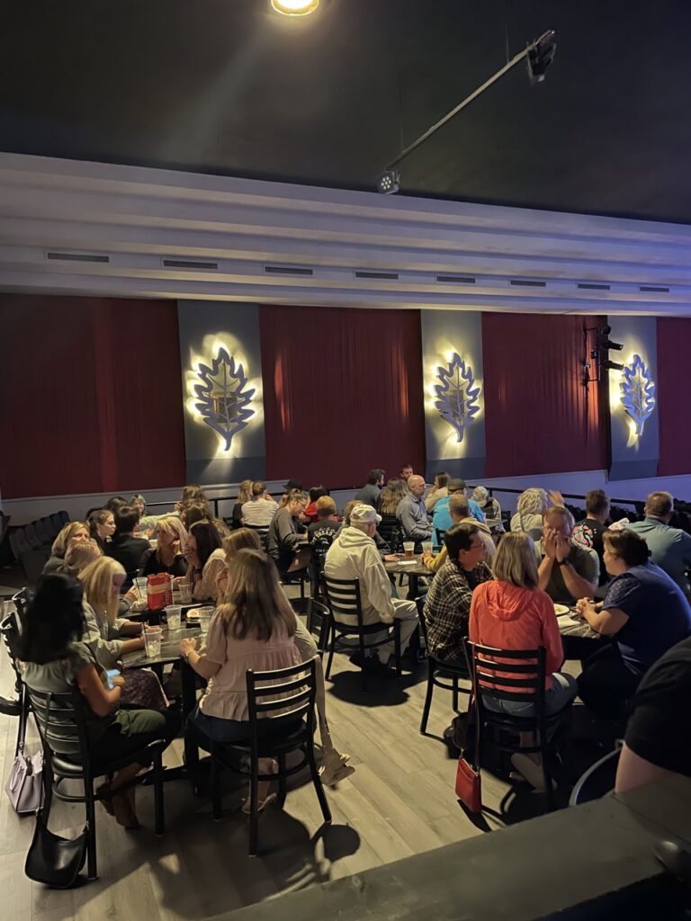 Groups of people sit at tables in the Oaks Theater.