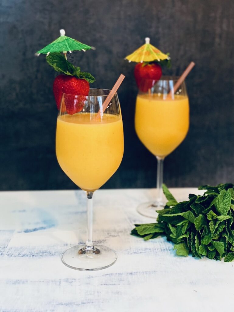 Two tall glasses with stems hold Tropical Mango Smoothie with strawberries on the rim with little cocktail umbrellas in the strawberries.