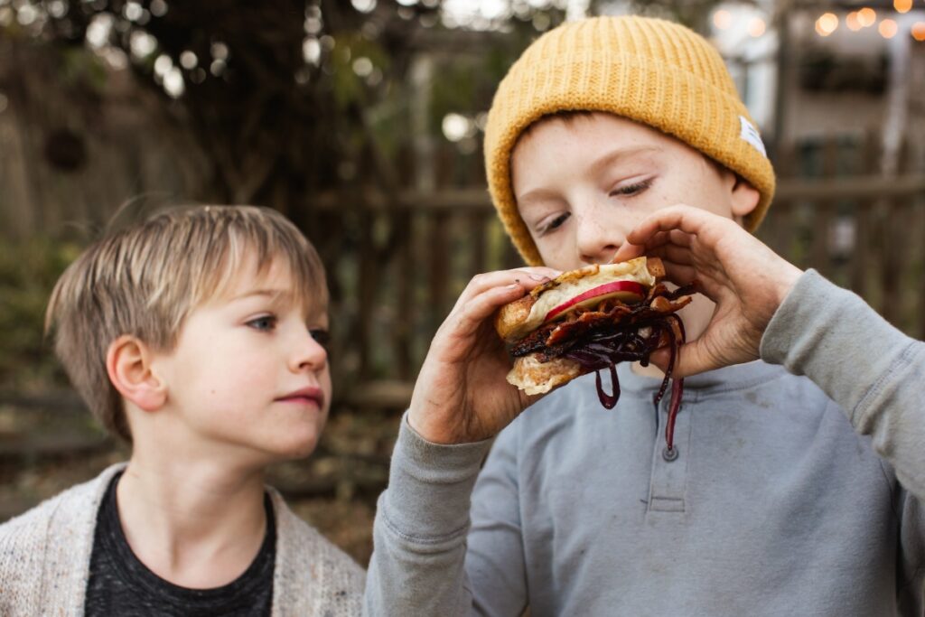A young boy in a yellow beanie takes a large bite of a gourmet grilled cheese sandwich featuring bacon, apple, and caramelized onions during an outdoor autumn gathering.