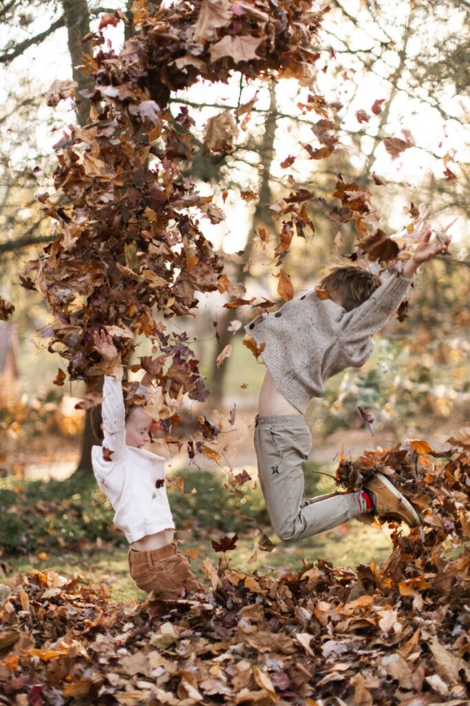 Two young children jumping into a large pile of autumn leaves and throwing them into the air during an outdoor family gathering.