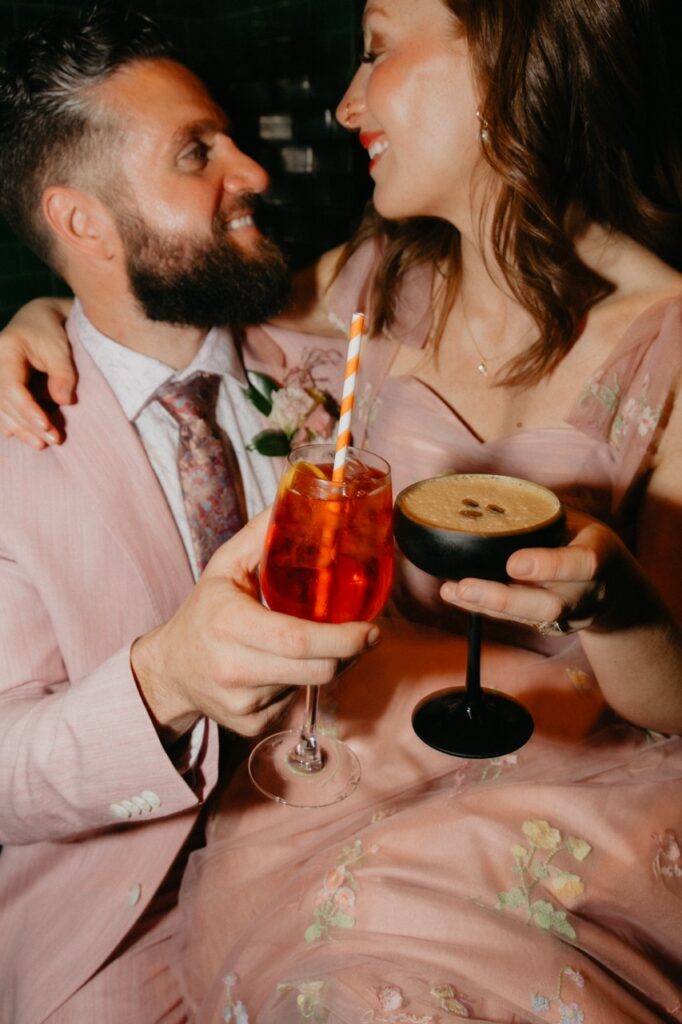 A woman and man in a pink dress and suit hold an aperol spritz and espresso martini.