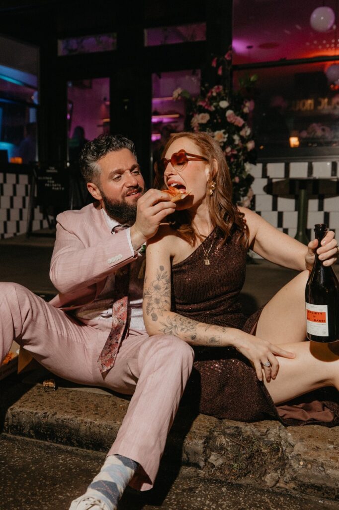 A man in a pink suit feeds his wife in a dress pizza.
