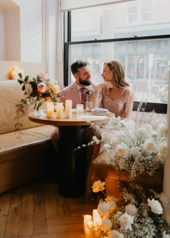 A couple sits at a small round table surrounded by white and pink wedding florals.