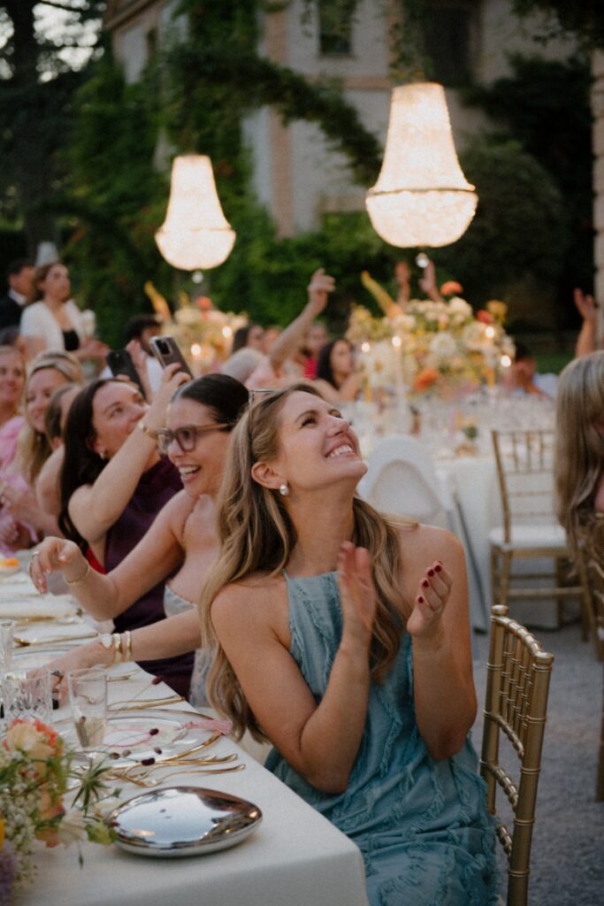 Wedding guests sitting at a long dinner table under glowing crystal chandeliers during an outdoor reception in the garden of Relais Villa Corallo in Italy.