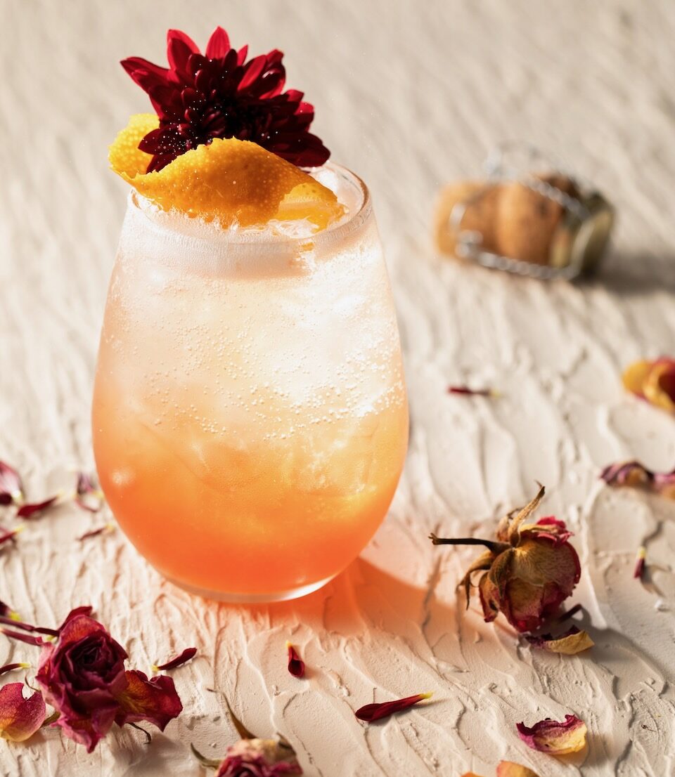 A short glass holds a When Harry Met Sally inspired spritz with an orange peel and edible flower garnish as a champagne cork and rose petals scatter around the glass.