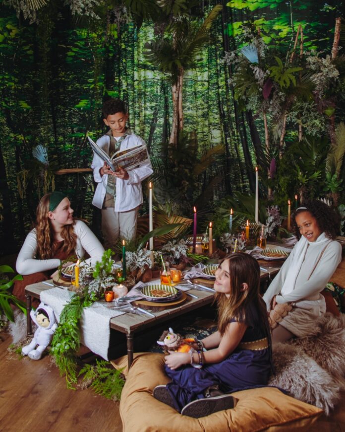 Children at a "Where the Wild Things Are" themed birthday party sitting on plush floor pillows around a rustic driftwood table in a mystical forest setting.