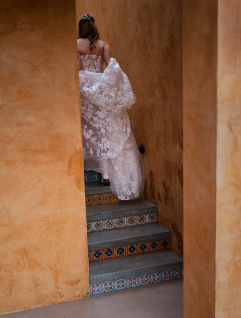 A bride in a floral lace wedding gown ascending a decorative tiled staircase at Villa Santa Cruz in Todos Santos, Mexico.