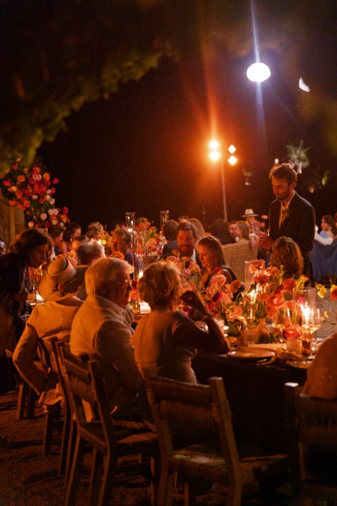 A candlelit outdoor wedding reception dinner at night featuring long wooden tables, vibrant floral arrangements, and guests engaged in conversation under warm lighting.