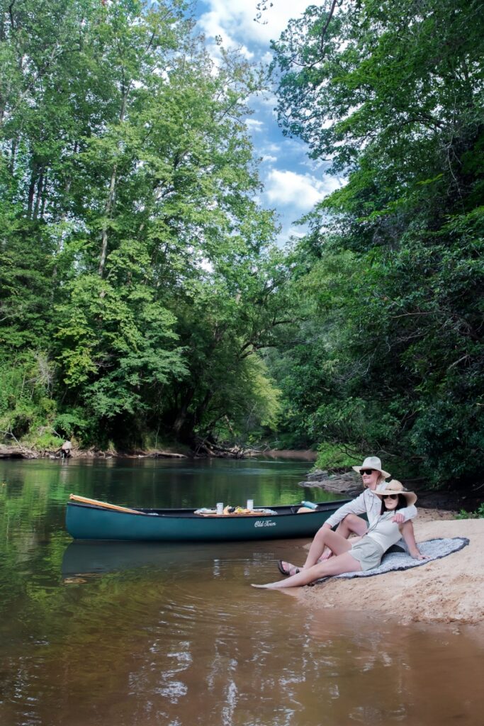 A couple wearing sun hats relaxes on a sandy riverbank next to their green canoe during a peaceful summer picnic in the Georgia highlands.