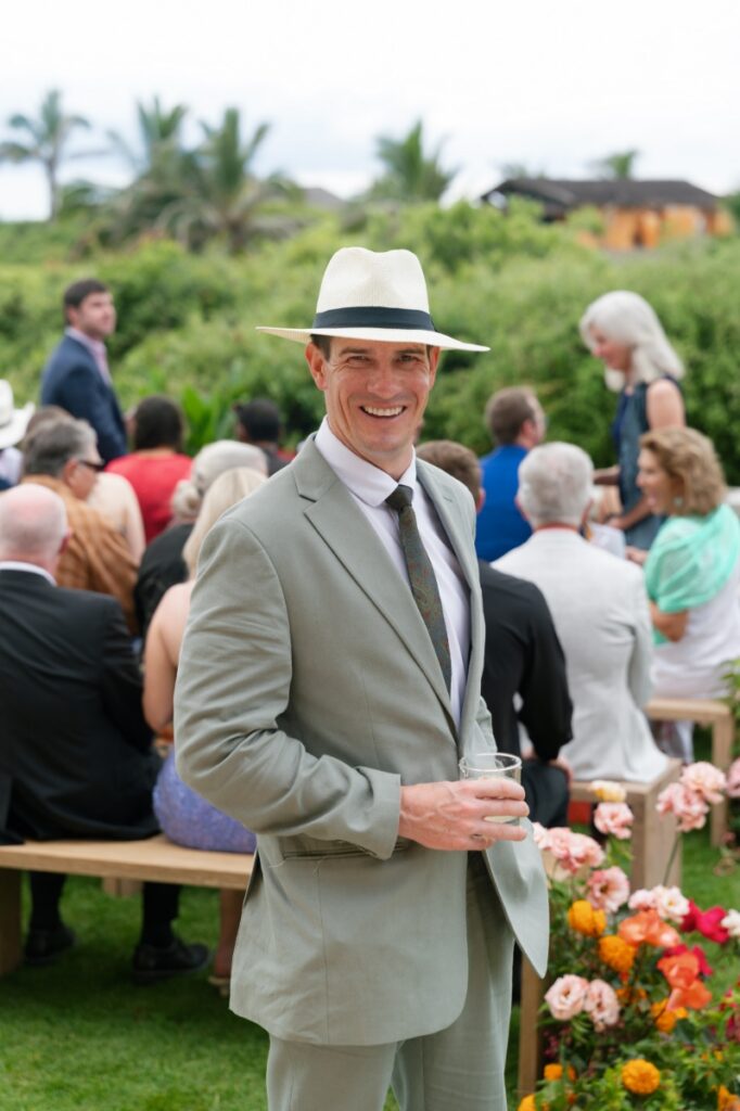 A wedding guest in a light grey suit and white straw hat smiling while holding a cocktail at an outdoor ceremony on the main villa lawn.