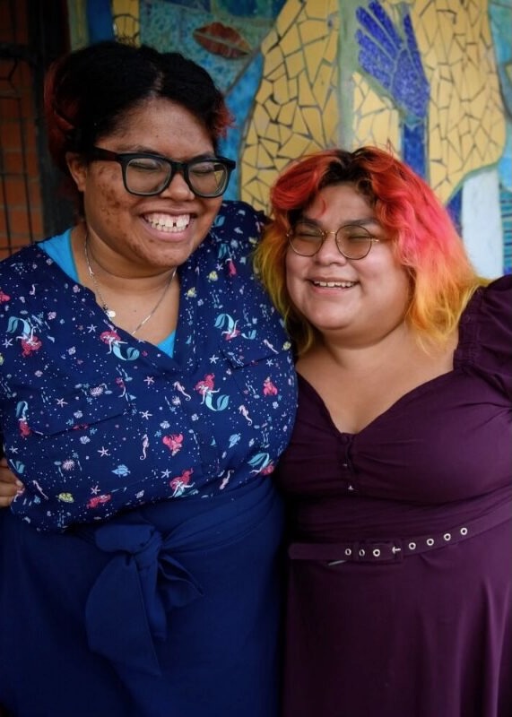 Two smiling women standing together in front of a colorful mosaic mural as part of a community human services initiative.