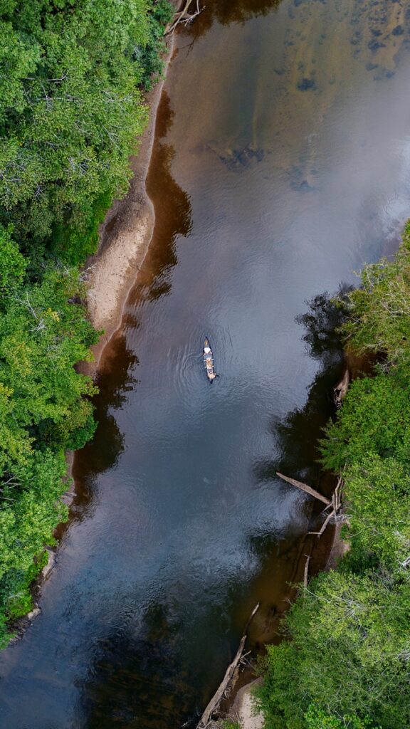 A high-altitude aerial view of a single canoe with two people paddling down a calm, winding river flanked by a dense green forest canopy in the Georgia highlands.
