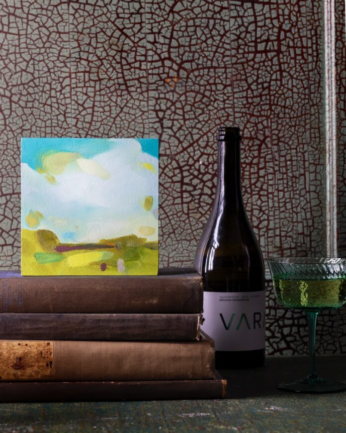 A small painting of a bright landscape by Nicole Renee Ryan sits on a stack of books beside a bottle of Vara wine.