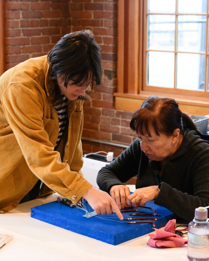 A woman at an art workshop shows another woman tips on a blue mat.