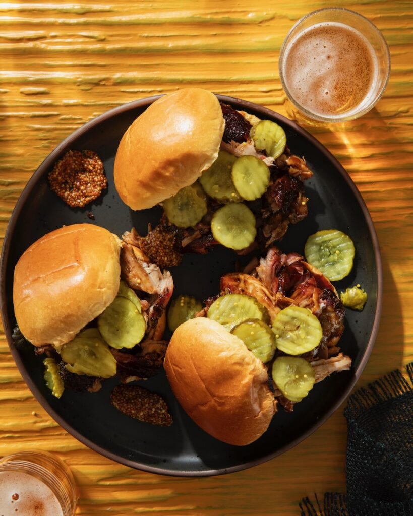 Three BBQ pork sandwiches with pickles on top of the meet and a beer in the top right corner.
