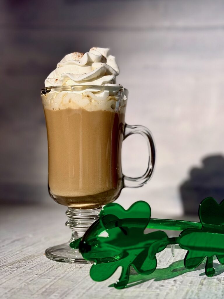 An Irish Coffee made with Baileys sits in a mug on a white table with whipped cream on top and a pair of shamrock sunglasses in front of the glass.