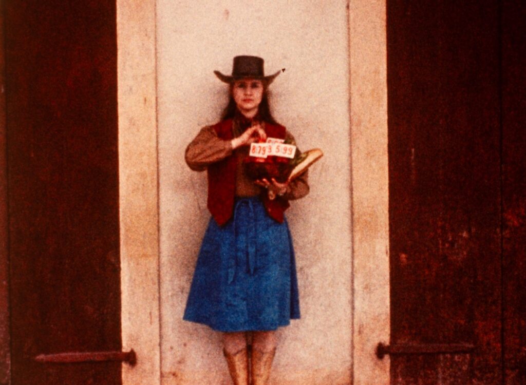 A woman stands in front of a beige background with a jean shirt, vest, and cowboy hat on.