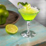 A green basil and cucumber gimlet in a Nick and Nora glass with half a lime sitting beside it.