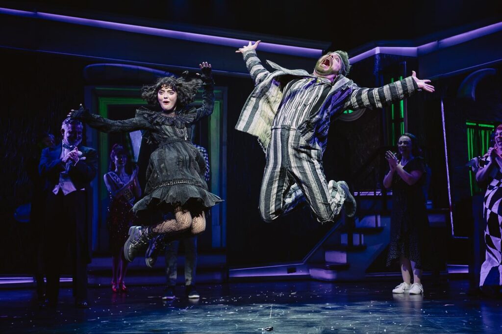 Beetlejuice and Lydia jump up in the air with their arms up, side by side.