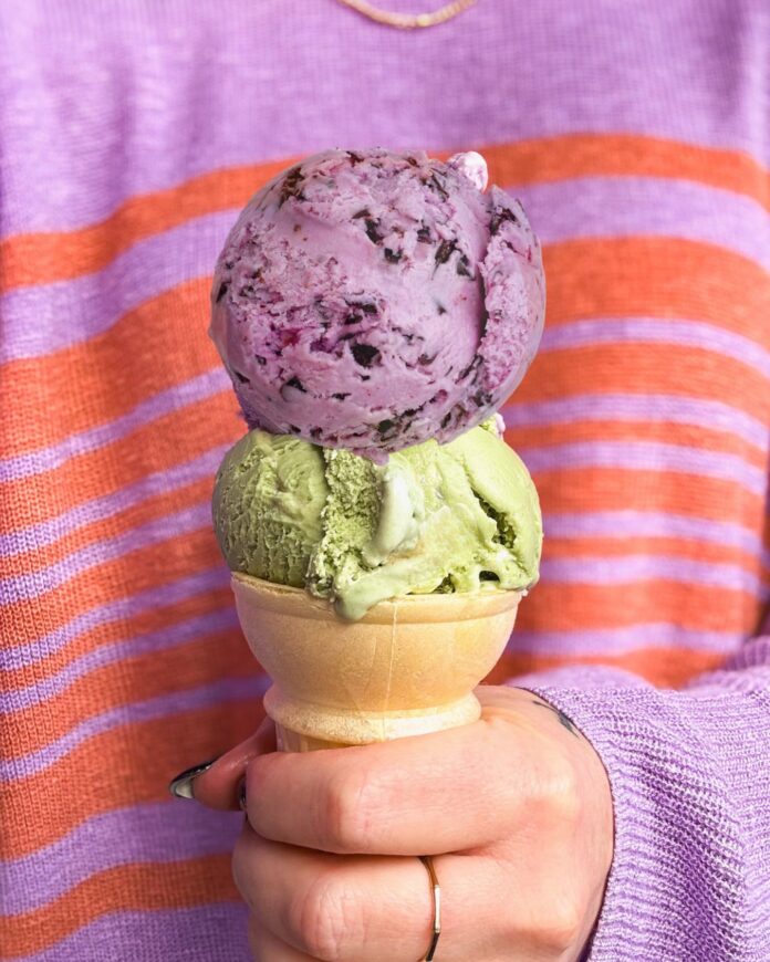 A person in a purple sweater holds a two scoop stack of ice cream in a cake cone.