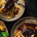 Two plates with Black Pepper Crusted Short Ribs, a corn hash, and grilled peppers in yellow and green.