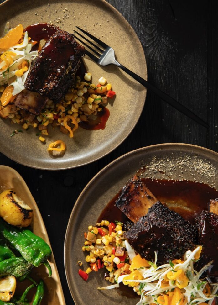 Two plates with Black Pepper Crusted Short Ribs, a corn hash, and grilled peppers in yellow and green.