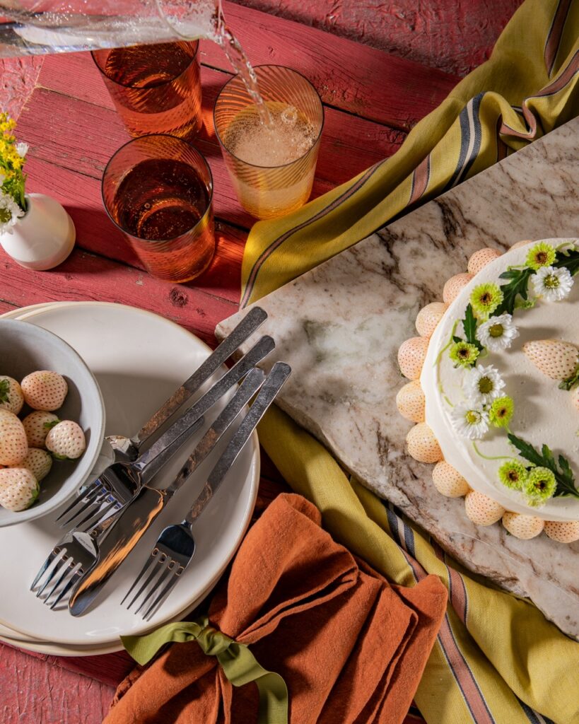 A stack of white plates with forks and white strawberries sits below three glasses and to the left of a white cake on a piece of stone.