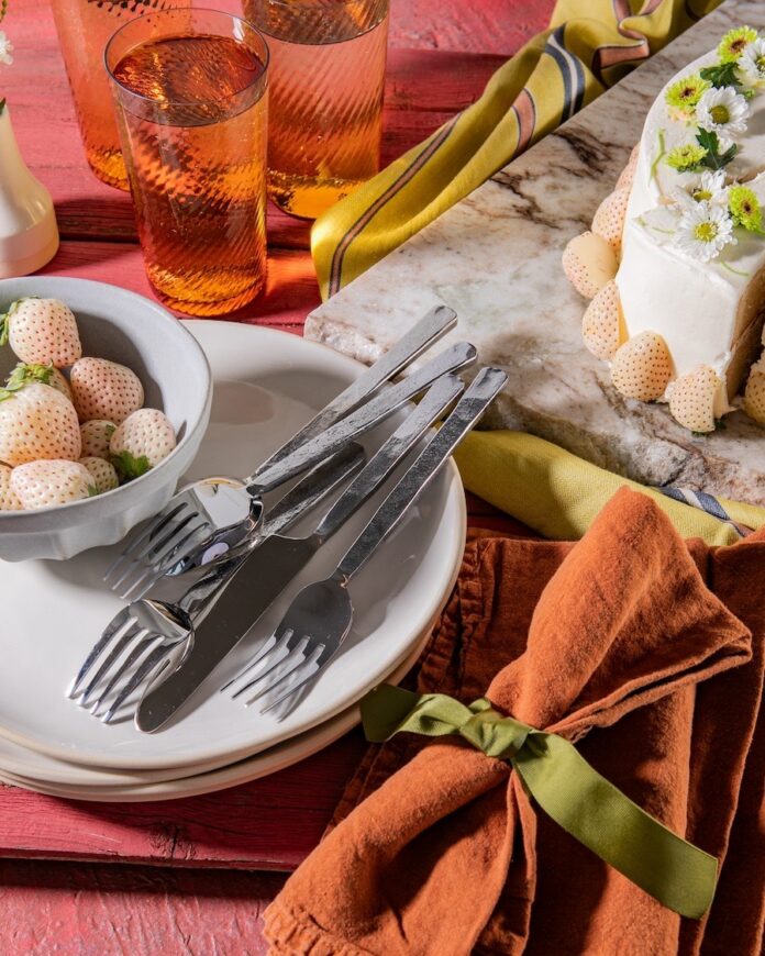 A stack of white plates beside an orange napkin, below glasses of water and a white strawberry cake.